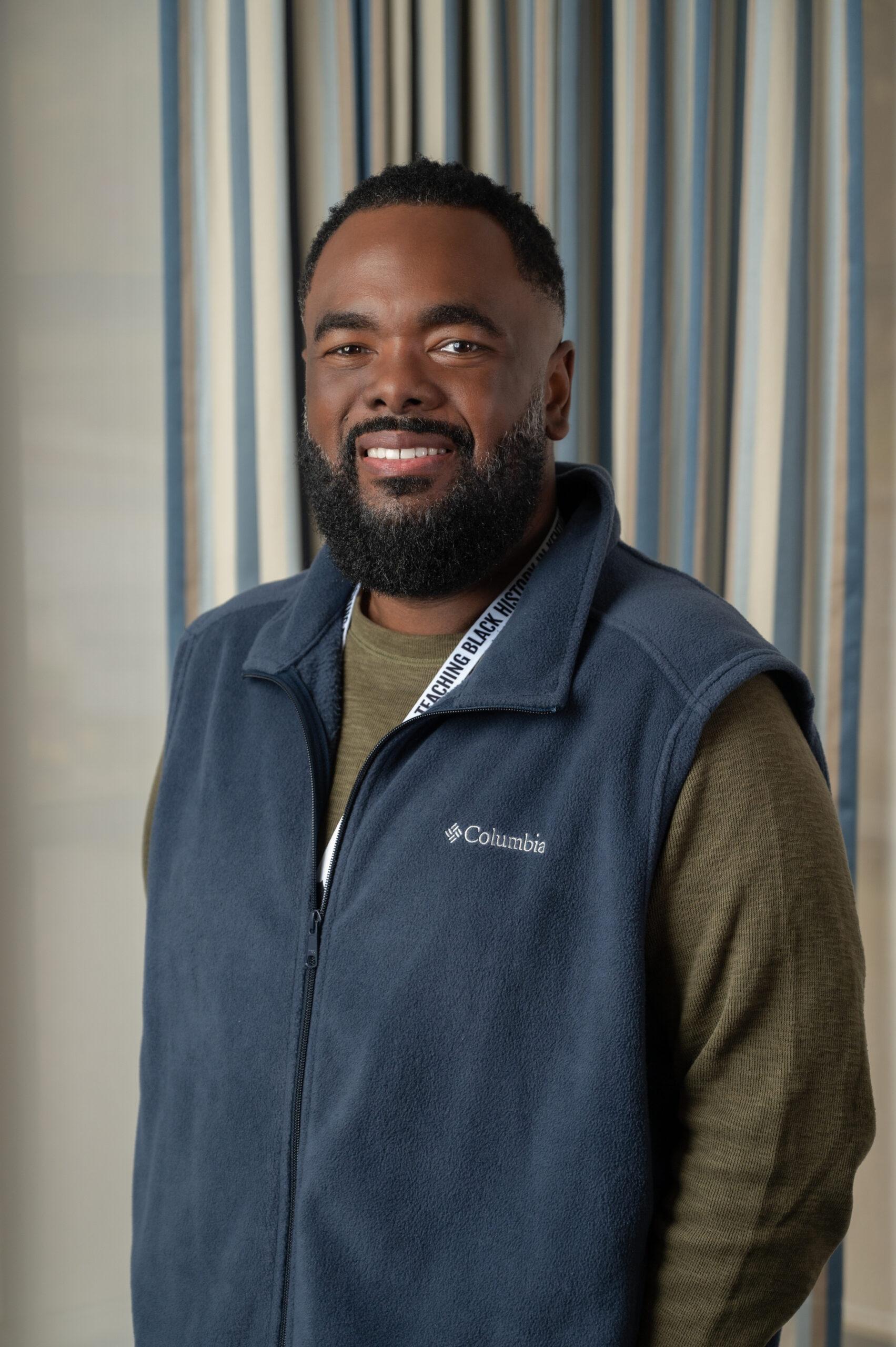 Chaka Cummings stands in front of a gray background. She hs smiling at the camera and is wearing a a blue sweater vest and green long sleeve shirt underneath.
